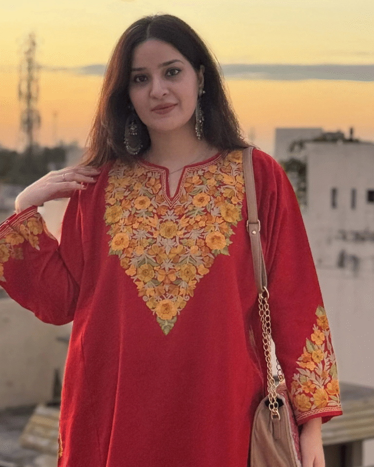 A woman posing outdoors at sunset, wearing a deep red Kashmiri Phiran featuring dense, yellow and gold floral Aari or Tilla embroidery across the yoke and cuffs.