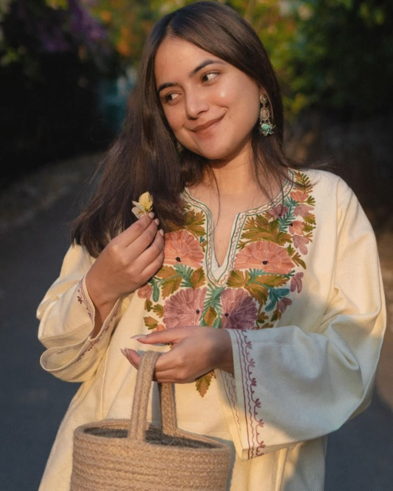 A woman smiling outdoors, holding a yellow flower, wearing a cream linen or cotton tunic featuring soft, pastel-colored Aari floral embroidery on the yoke, carrying a woven straw bag.