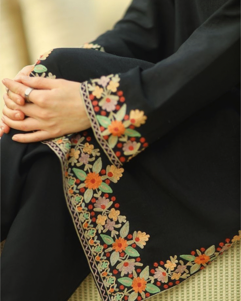 Close-up of a person wearing a black kurta, showing intricate Kashmiri Aari or crewel floral embroidery along the kurta hem and sleeve cuffs in orange, green, and pink colors.